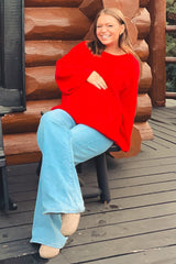 woman wearing a red oversized maternity sweater and light blue flared jeans, sitting and holding her baby bump