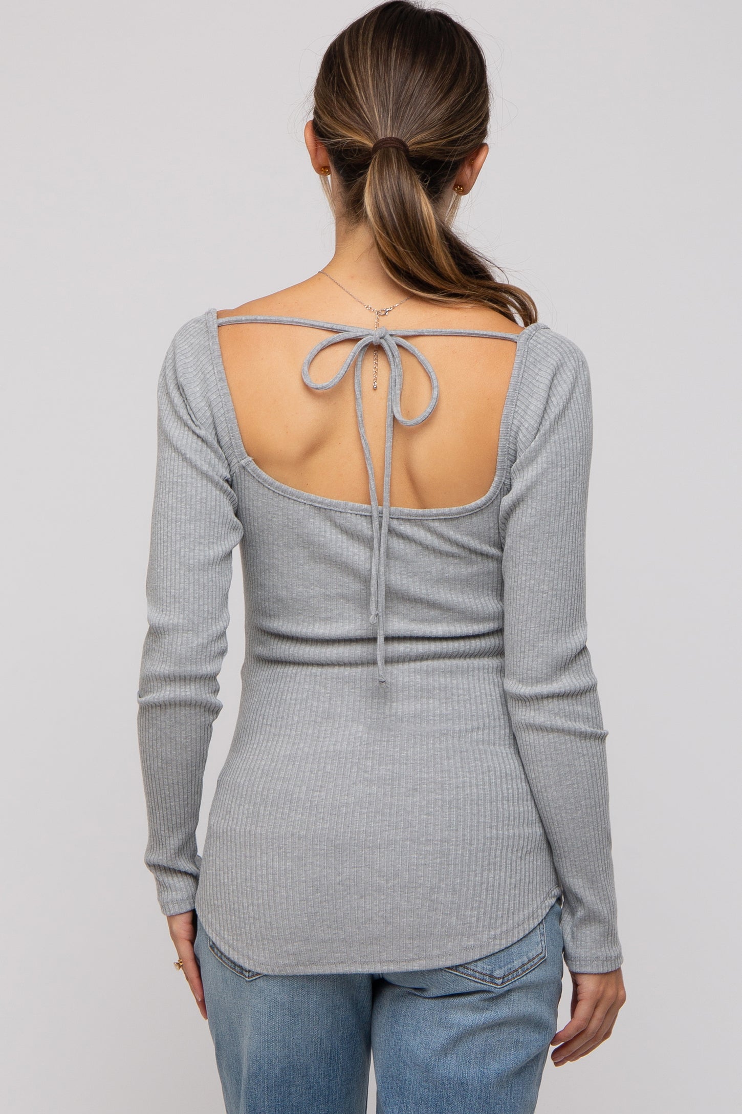 woman wearing a grey long sleeve maternity top with a cut-out back and tie detail, paired with distressed jeans