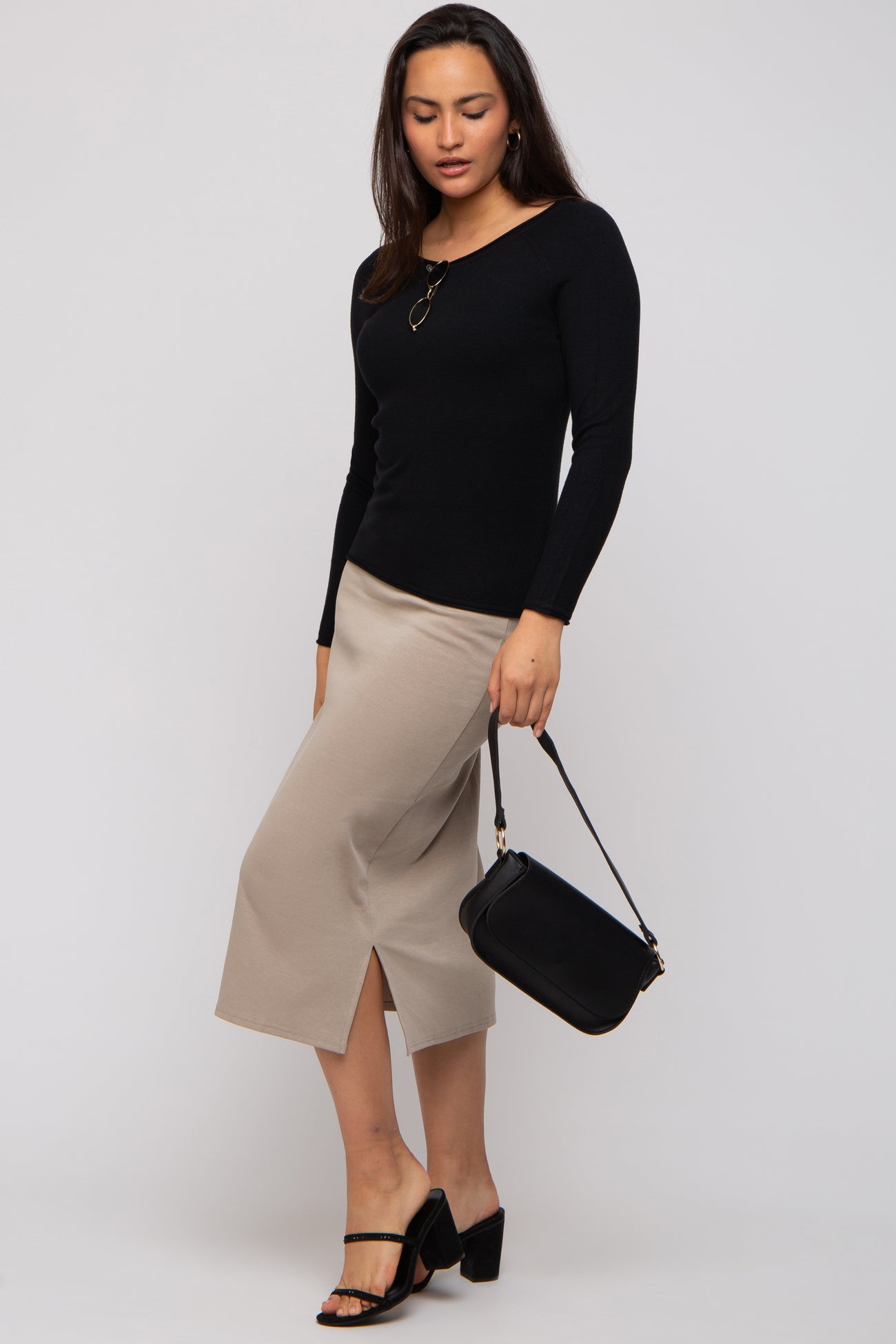 woman wearing a black long sleeve top and a beige midi skirt, holding a small black handbag, with strappy heels
