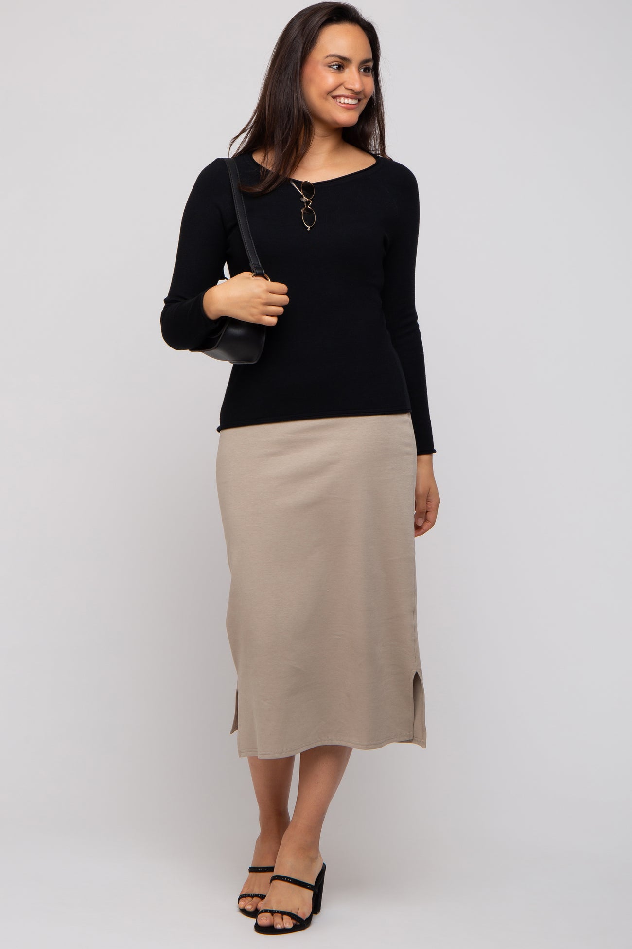 woman wearing a black long sleeve top and a beige midi skirt with slits, posing with a black shoulder bag
