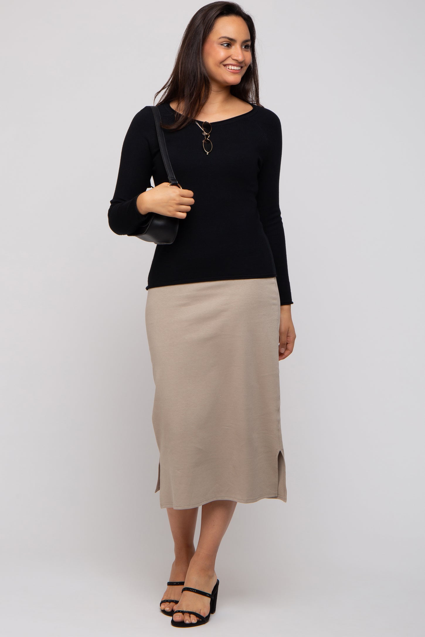 woman wearing a black long sleeve top and a beige midi skirt with slits, posing with a black shoulder bag