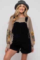 woman wearing a beige long sleeve top with patterned sleeves and black corduroy maternity overalls, smiling and posing