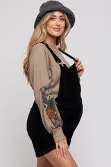 woman wearing a black corduroy maternity overall dress with a beige long sleeve top and a grey hat, showing her baby bump
