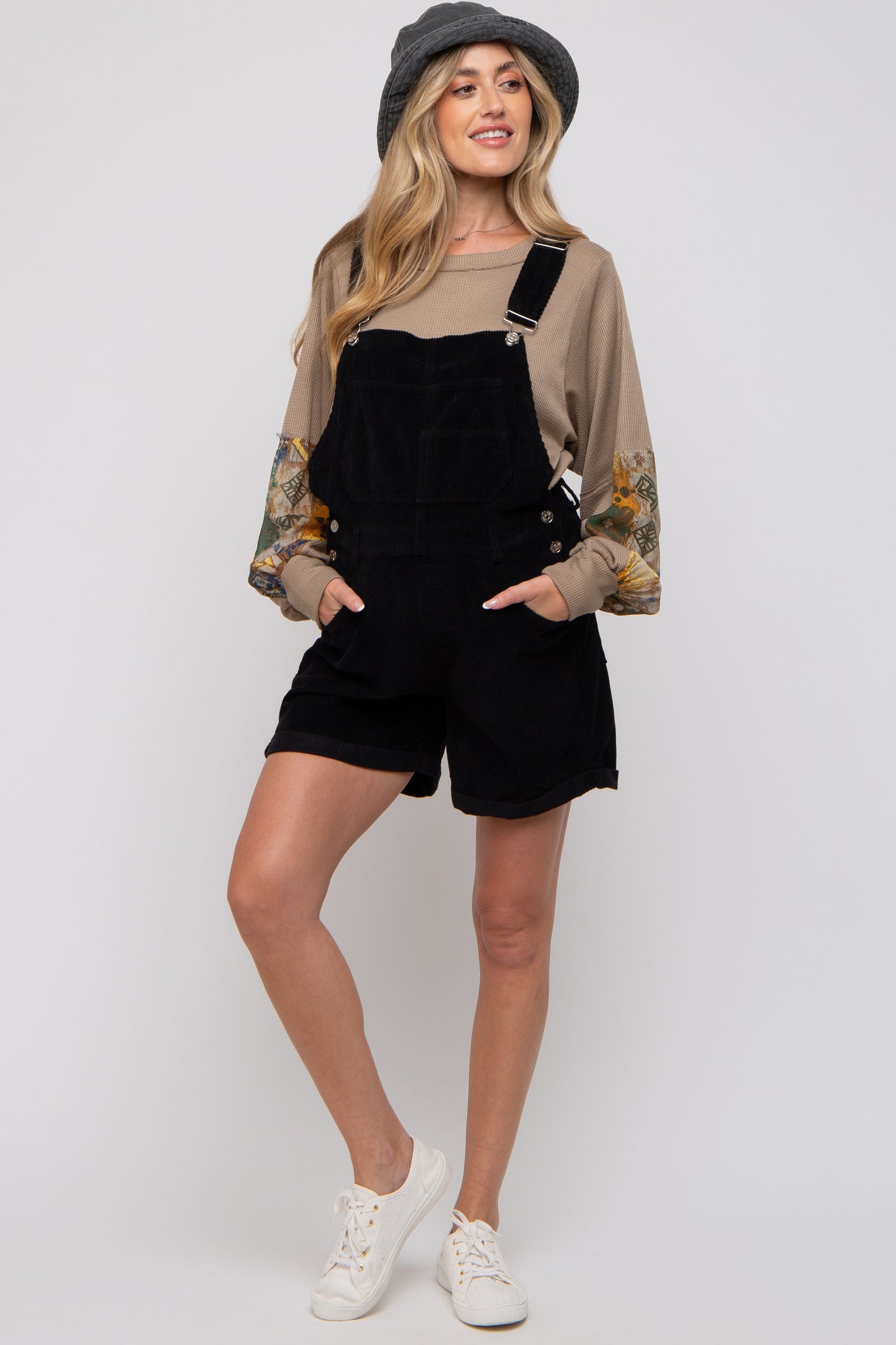 woman wearing a black corduroy maternity overall shorts with floral long sleeves and white sneakers
