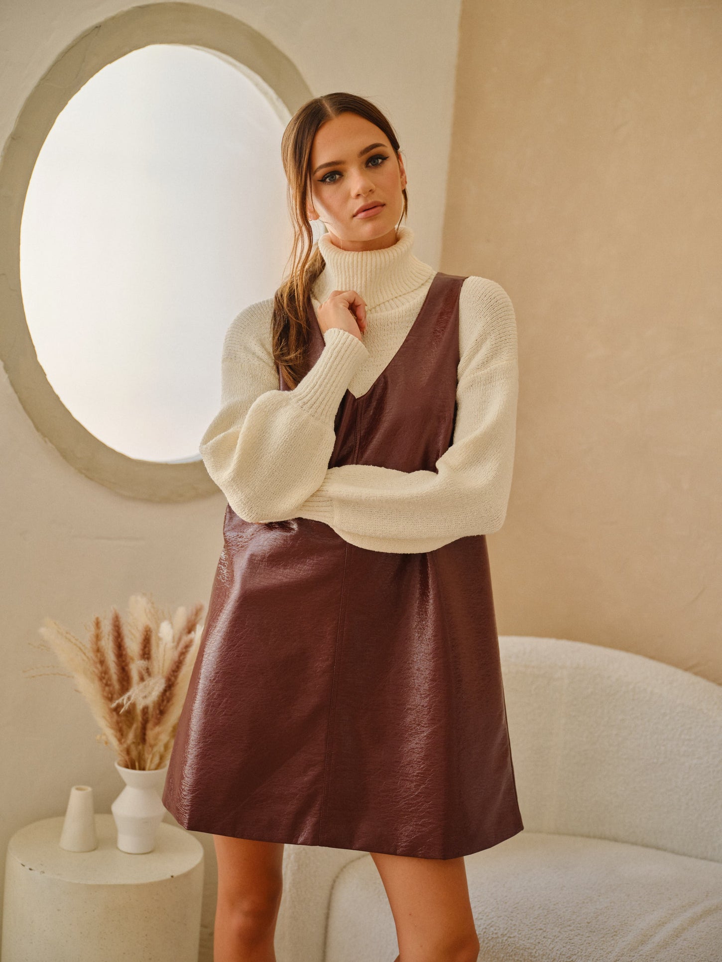 woman wearing a brown leather faux slip dress over a cream turtleneck sweater in a soft indoor setting