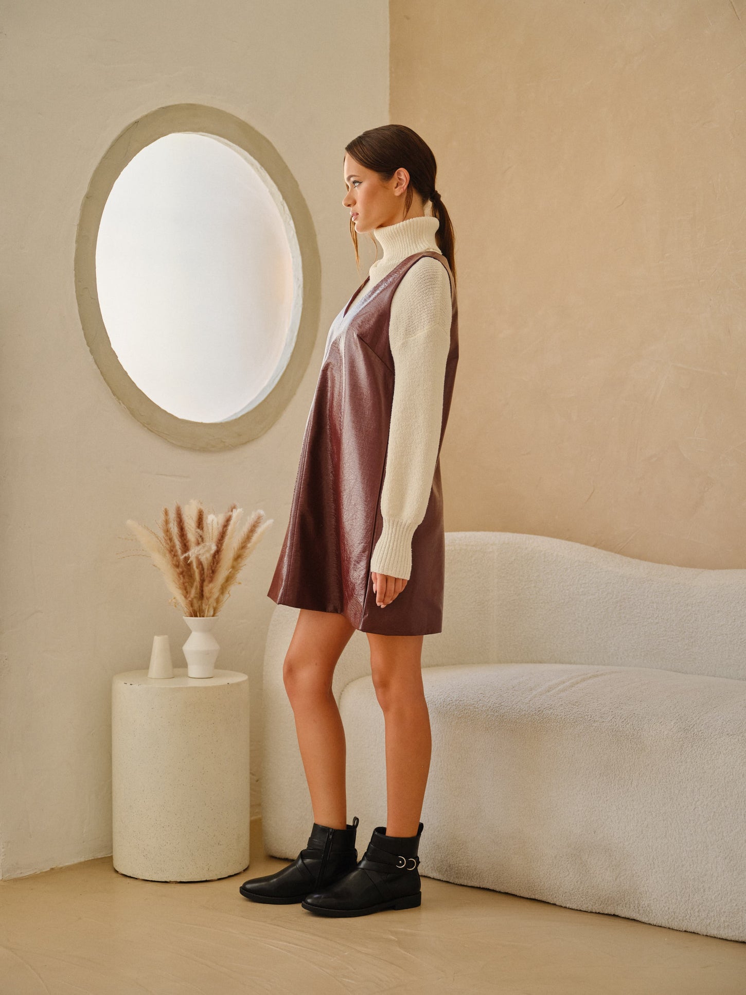 woman wearing a brown faux leather dress over a cream turtleneck sweater, standing beside a round window