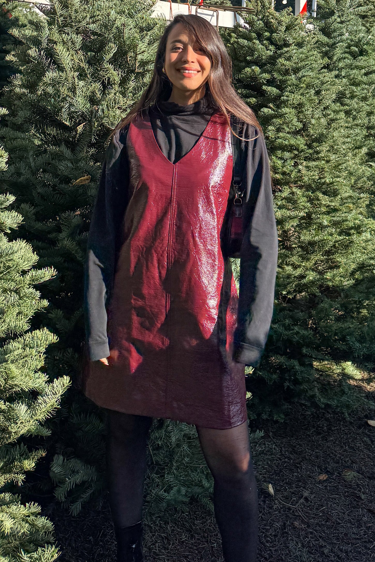woman wearing a burgundy faux leather sleeveless dress over a black turtleneck, surrounded by evergreen trees