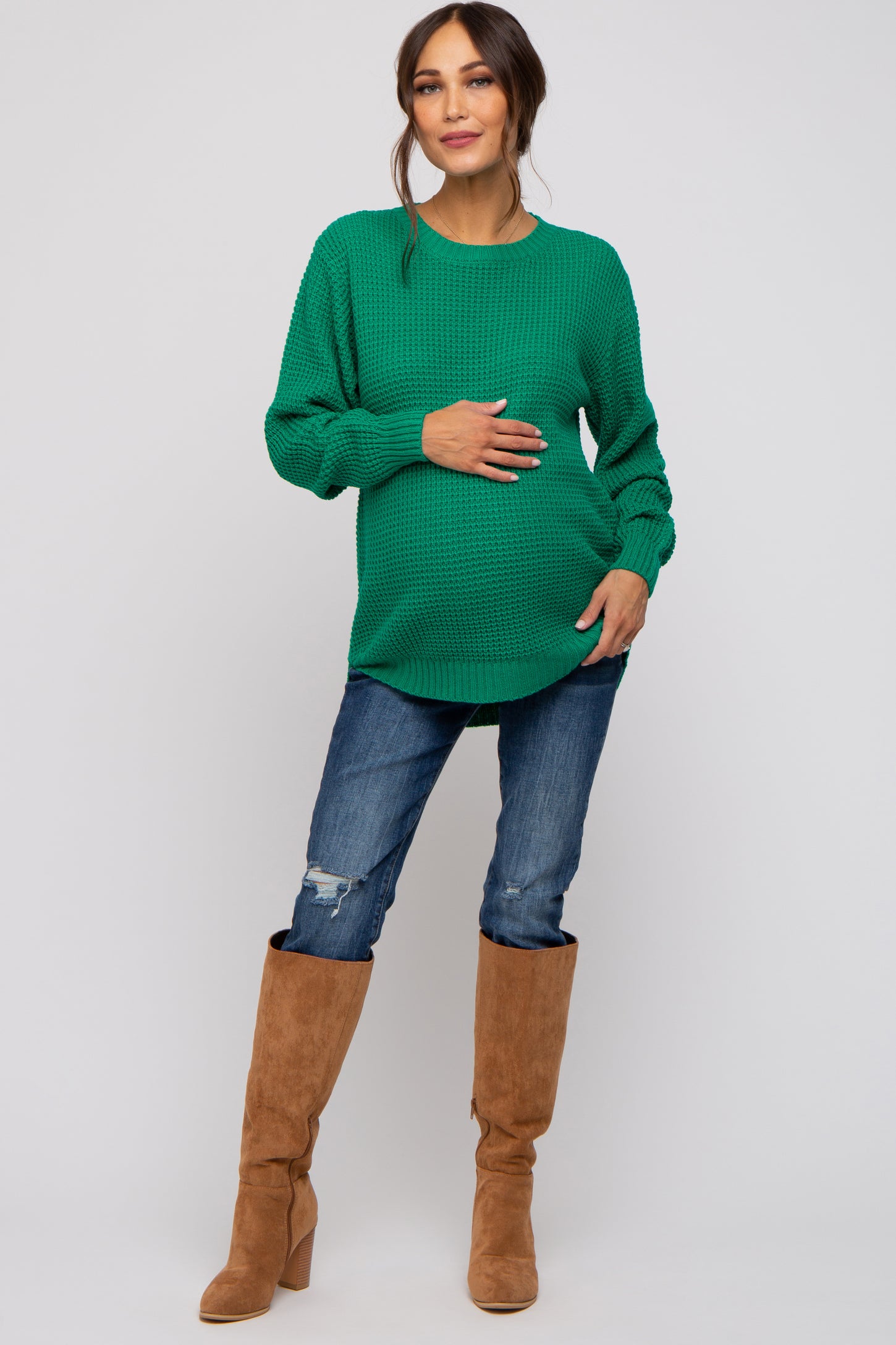 woman wearing a green oversized maternity sweater and holding her baby bump, paired with distressed jeans and knee-high boots