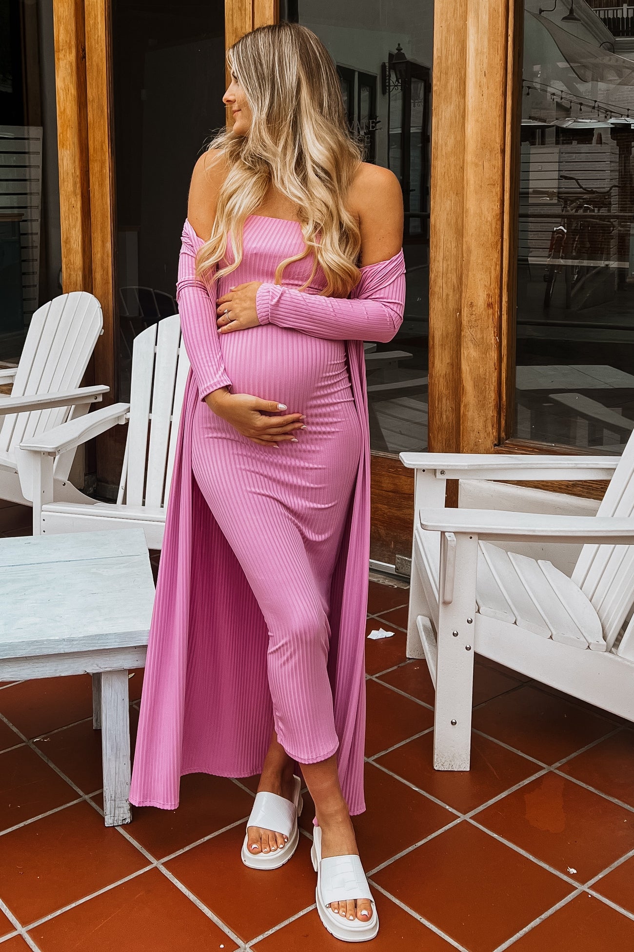 woman wearing a pink ribbed off-shoulder maternity dress with a long duster, holding her baby bump