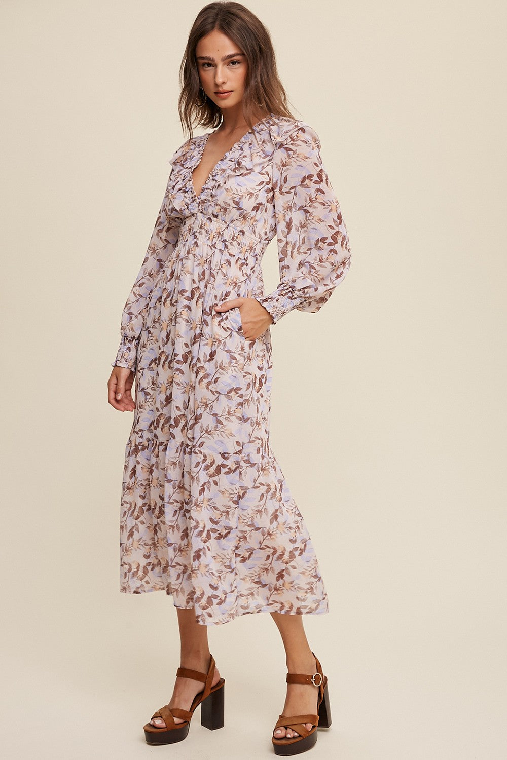 woman wearing a light purple floral maxi dress with long sleeves and ruffled neckline, standing in a neutral background