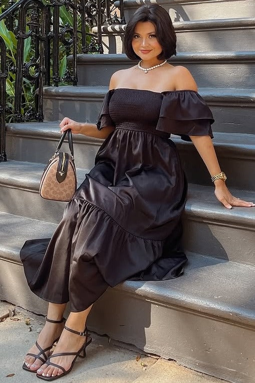 woman wearing a black off-the-shoulder maxi dress with ruffled sleeves and a tiered skirt, holding a small handbag