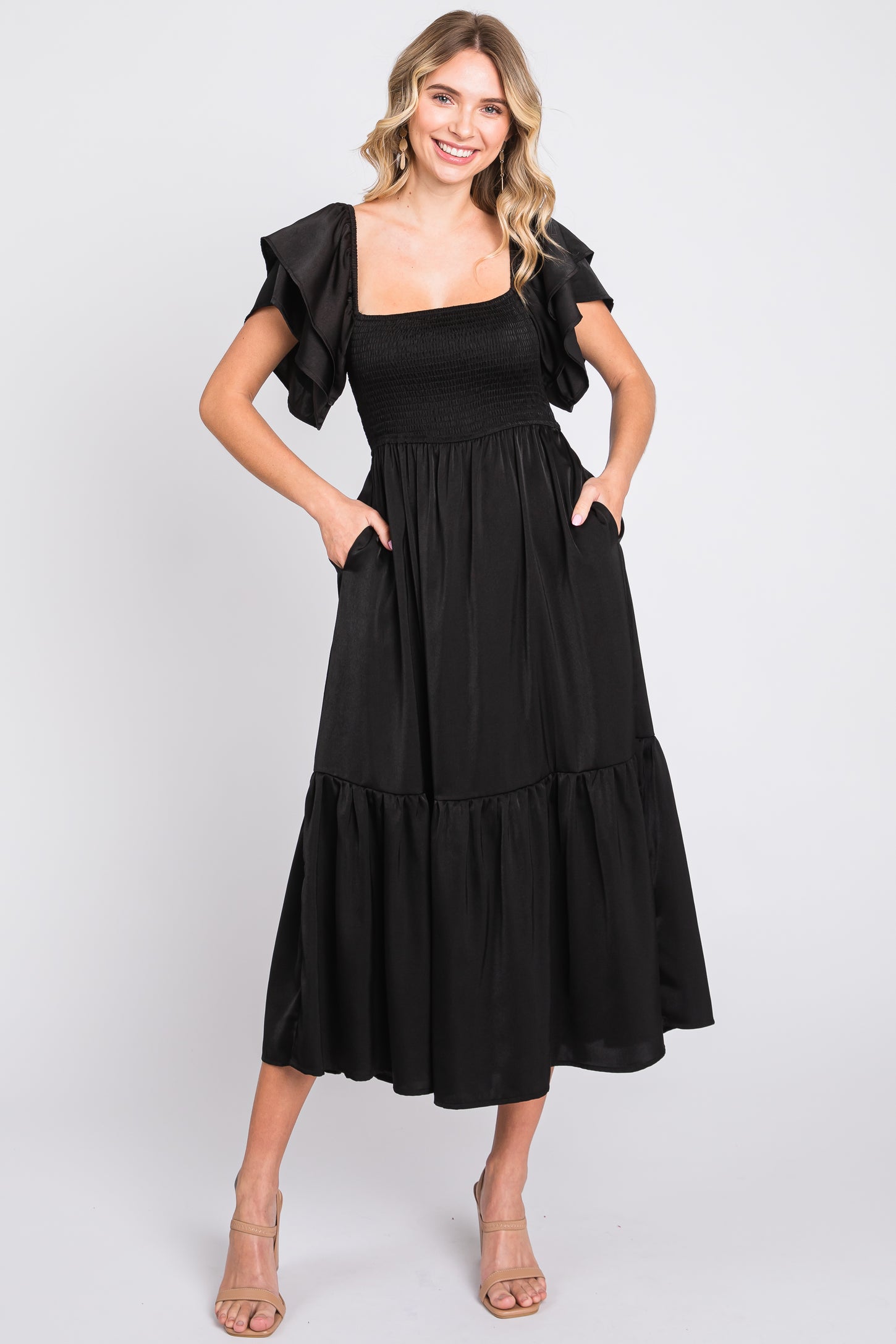 woman wearing a black maxi dress with ruffled sleeves and pockets, smiling and posing against a white background