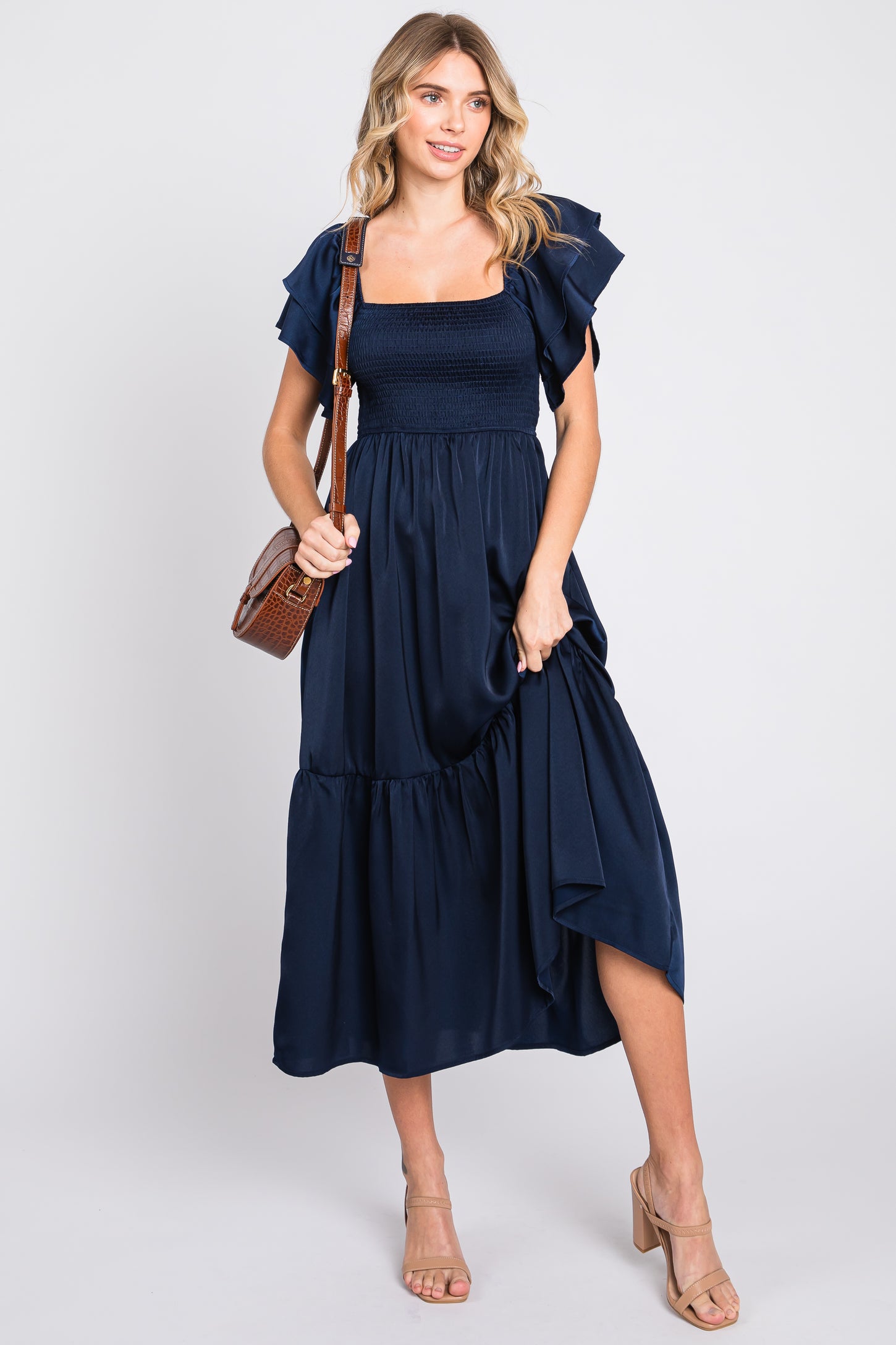 woman wearing a navy blue smocked maxi dress with ruffled sleeves, holding a brown handbag and wearing nude heeled sandals
