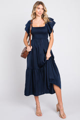 woman wearing a navy blue smocked maxi dress with ruffled sleeves, holding a brown handbag and wearing nude heeled sandals