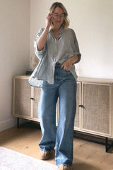 woman wearing a loose striped button-up shirt and high-waisted blue jeans, standing in a cozy room