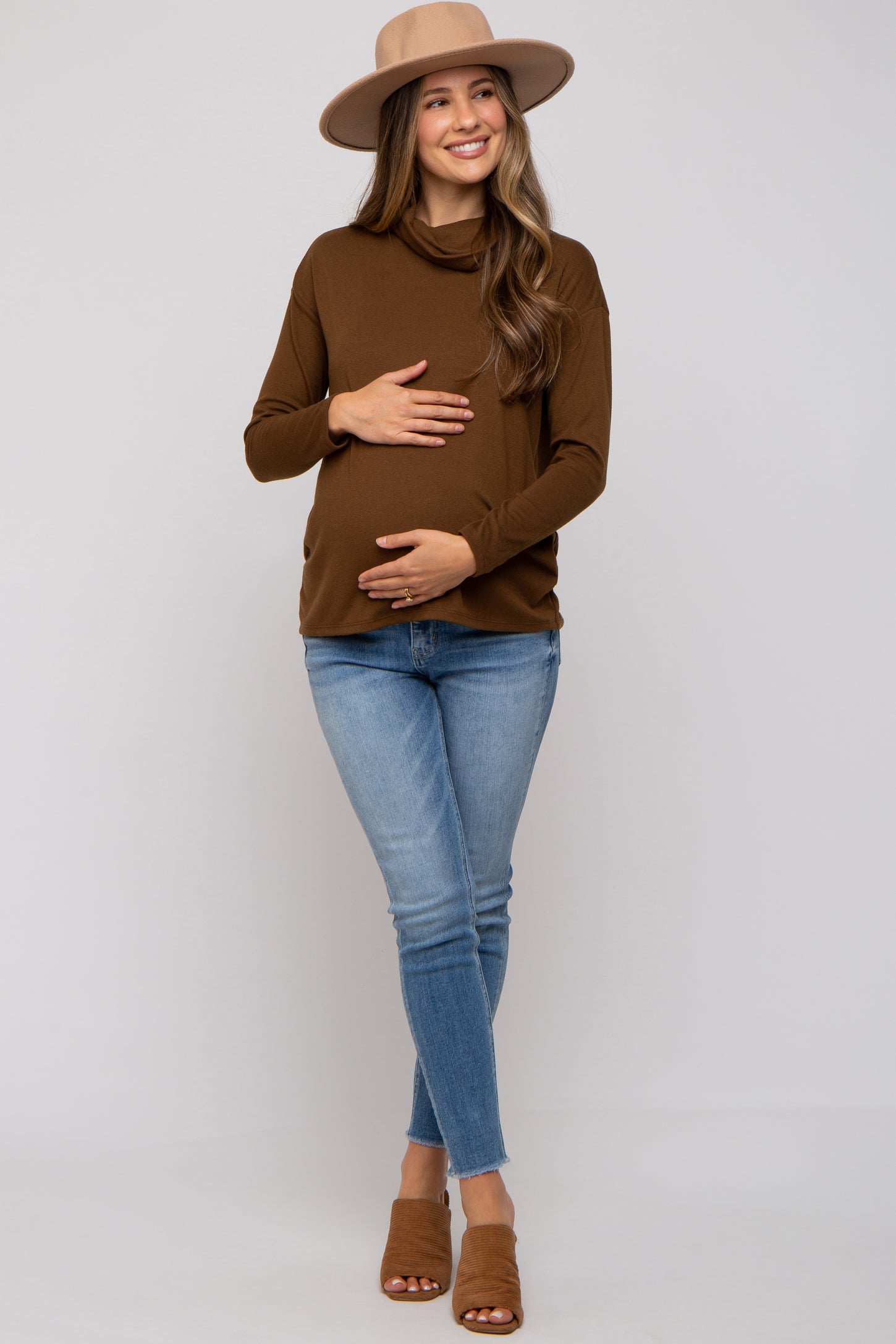 woman wearing a brown long sleeve maternity top and blue skinny jeans, holding her baby bump, wearing a hat and sandals