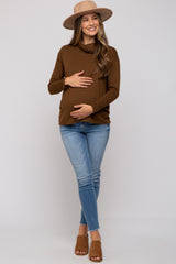woman wearing a brown long sleeve maternity top and blue skinny jeans, holding her baby bump, wearing a hat and sandals