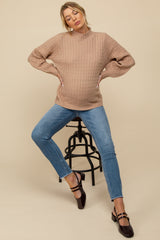 woman wearing a beige cable knit maternity sweater and blue skinny jeans, sitting on a stool with a visible baby bump