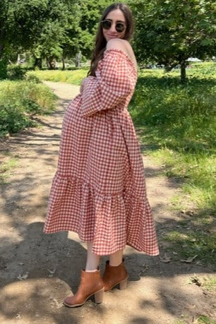 woman wearing a red and white gingham off-shoulder maternity dress with a ruffled hem and holding her baby bump