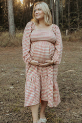 woman wearing a pink and white gingham maternity dress with long sleeves, holding her baby bump in a forest setting