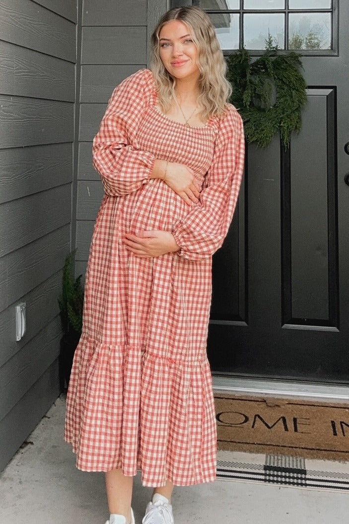 woman wearing a red and white gingham maternity dress with puff sleeves and tiered skirt, gently holding her baby bump