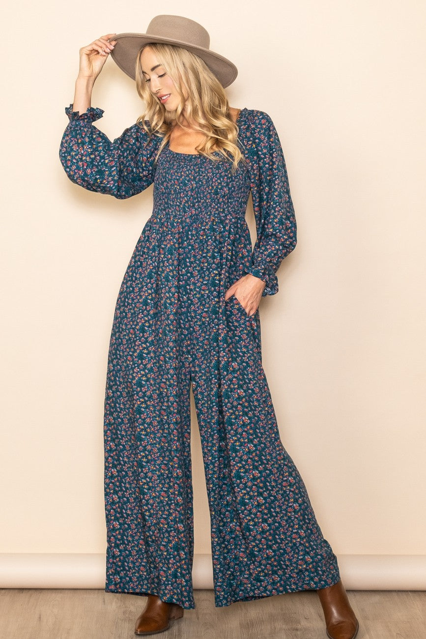 woman wearing a navy blue floral jumpsuit with long sleeves and wide legs, accessorized with a wide-brimmed hat