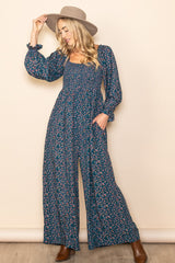 woman wearing a navy blue floral jumpsuit with long sleeves and wide legs, accessorized with a wide-brimmed hat