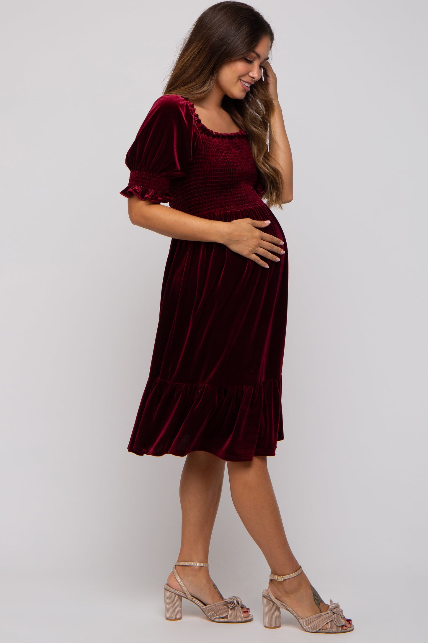 woman wearing a burgundy velvet maternity dress with short puff sleeves and a ruffled hem, gently cradling her baby bump