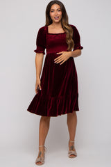 woman wearing a burgundy velvet maternity dress with short sleeves and a ruffled hem, holding her baby bump