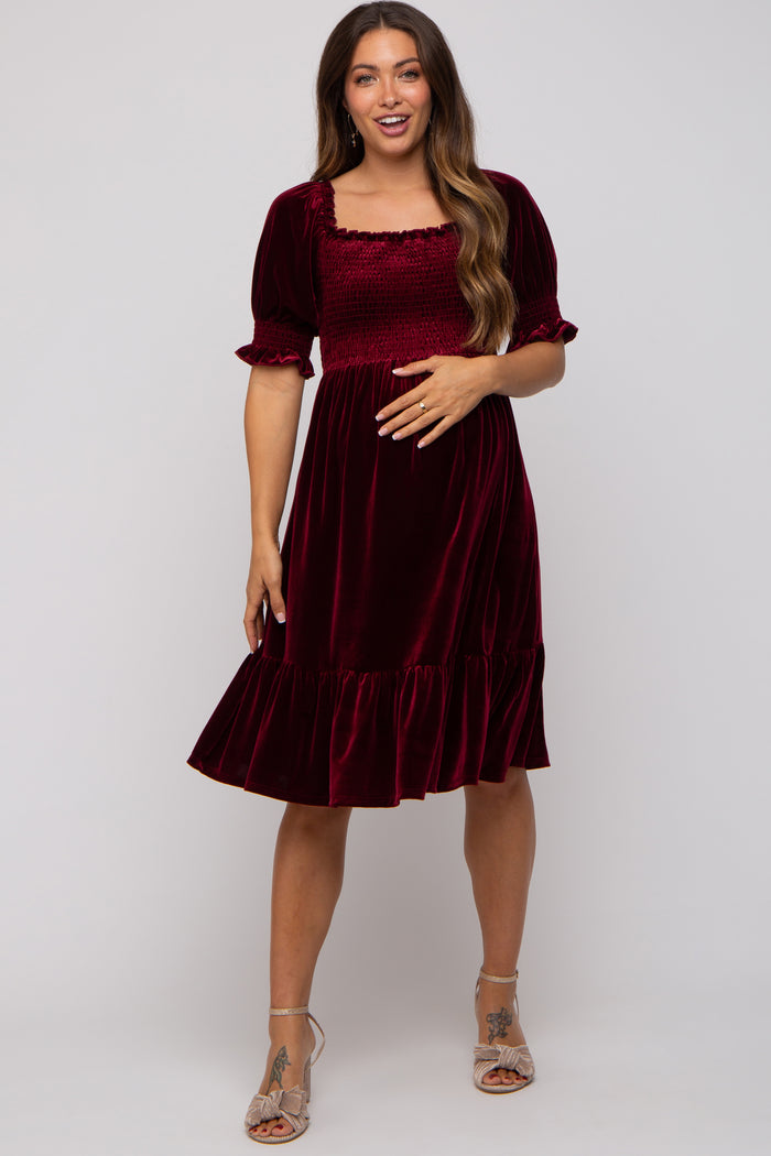 woman wearing a burgundy velvet maternity dress with short sleeves and a ruffled hem, holding her baby bump