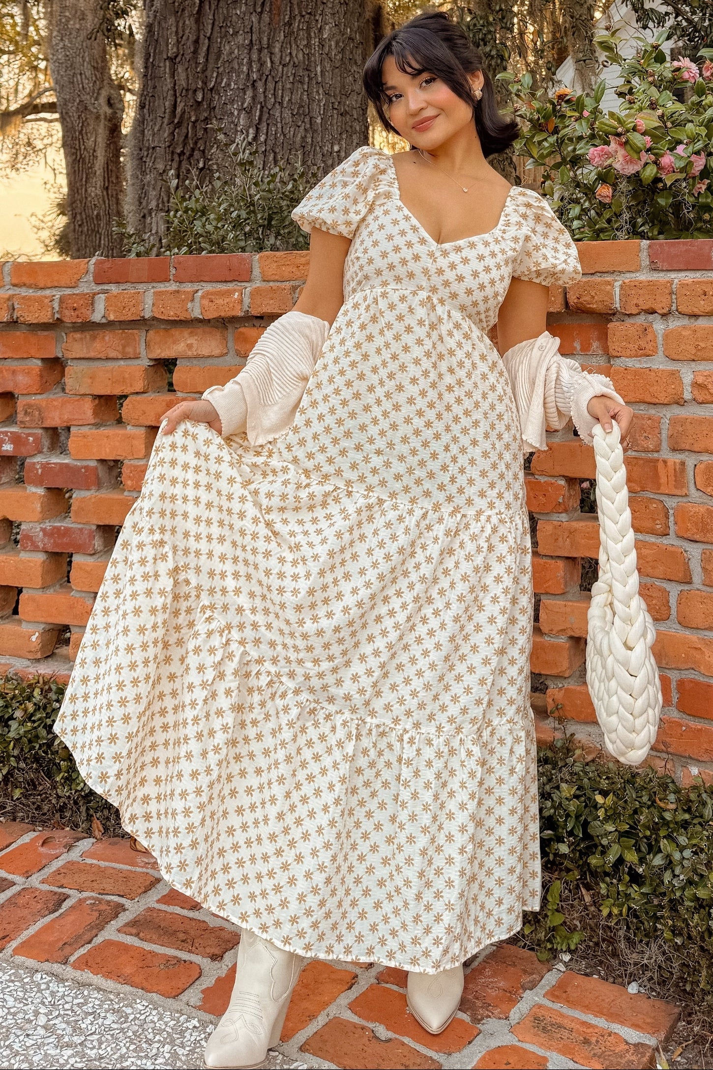 woman wearing a cream floral print maxi dress with puff sleeves and a tiered skirt, holding a braided handbag