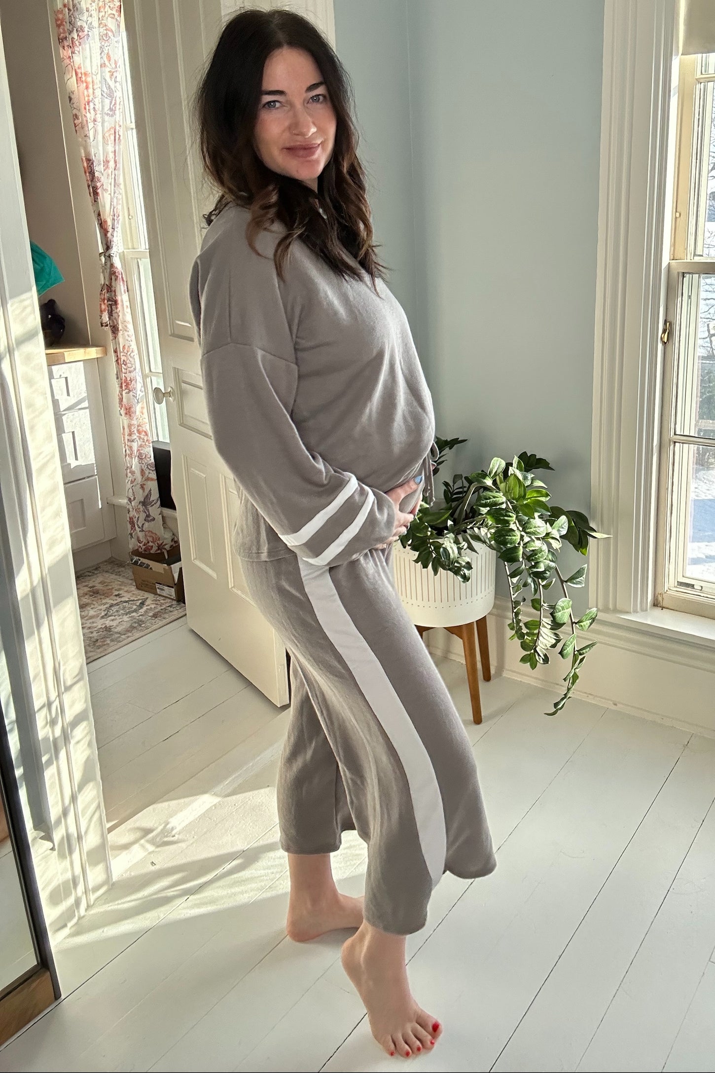 woman wearing a gray maternity lounge set with white stripes, showing her baby bump and standing beside a plant