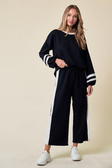 woman wearing a black lounge set with white stripes, featuring a loose top and wide-leg pants, standing against a neutral background