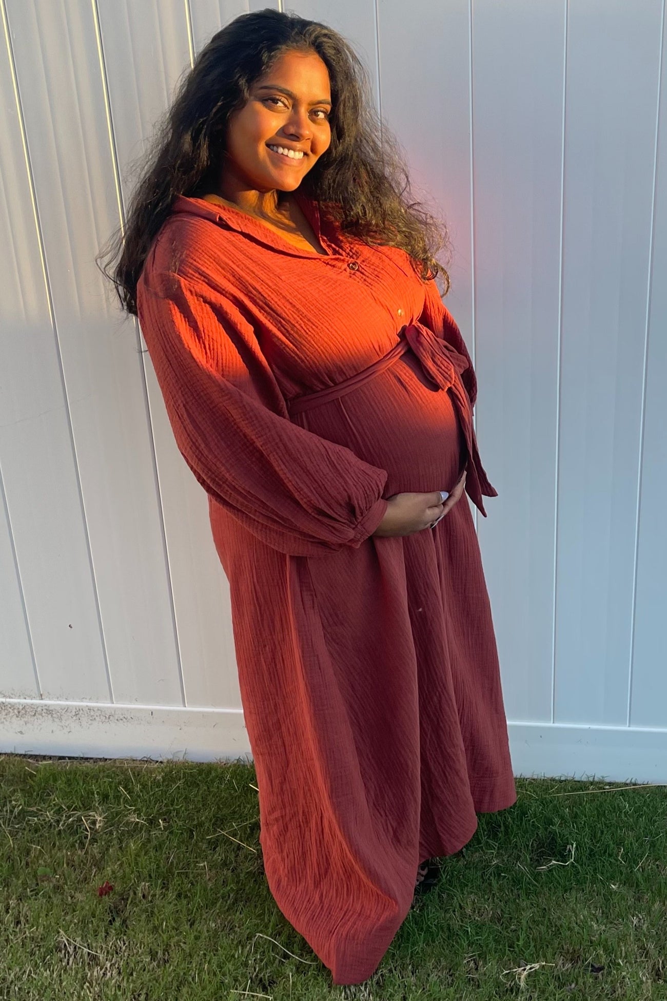 woman wearing a rust-colored maternity dress with long sleeves and a tie waist, smiling and showing her baby bump