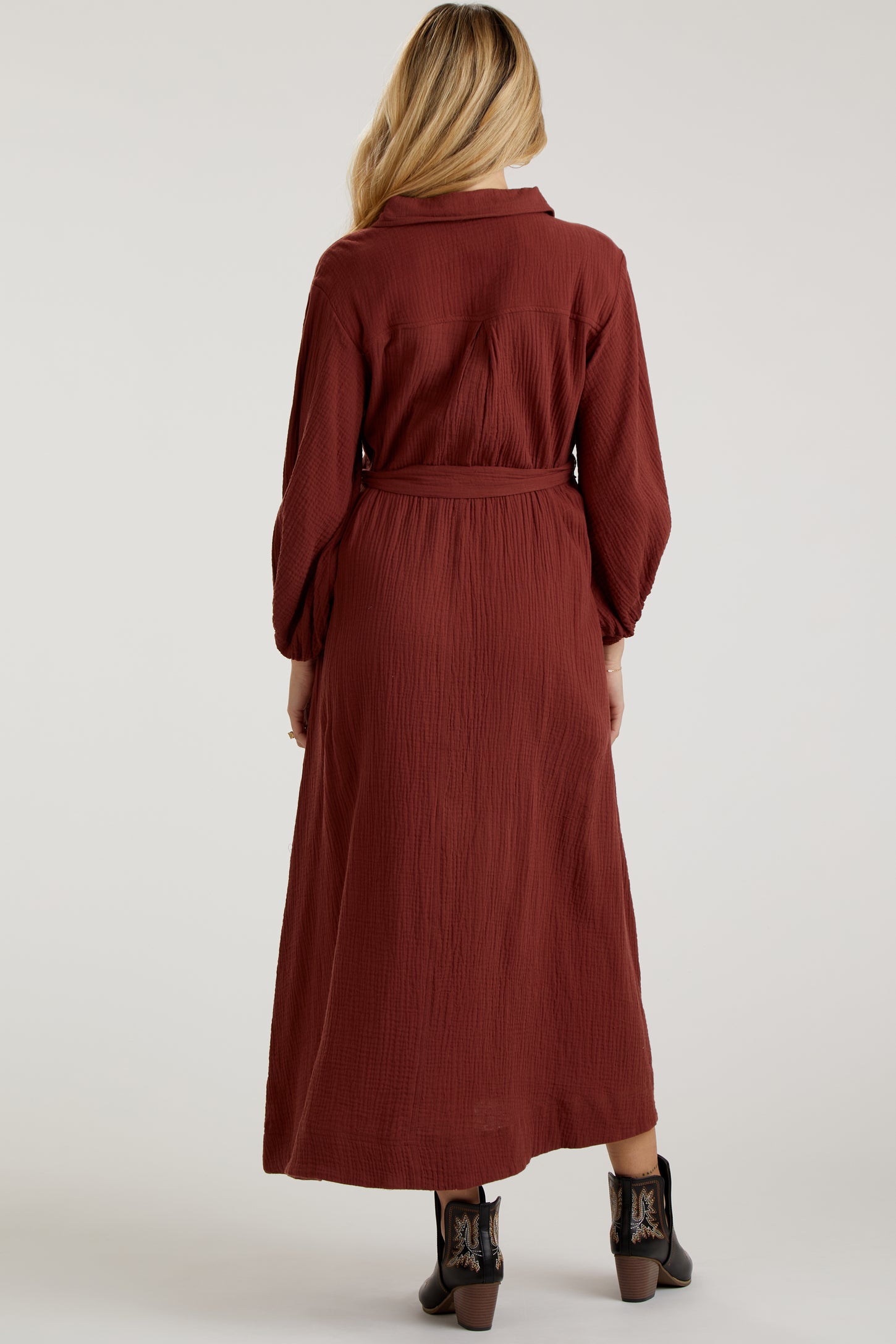 woman wearing a rust-colored long sleeve maternity dress with a belted waist and a flowing maxi length from the back