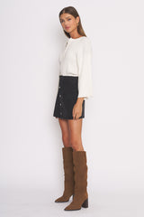 woman wearing a white long sleeve ribbed top, black button-front mini skirt, and tall brown suede boots