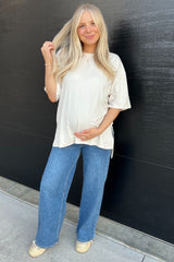woman wearing a cream oversized maternity top and blue wide-leg jeans, holding her baby bump
