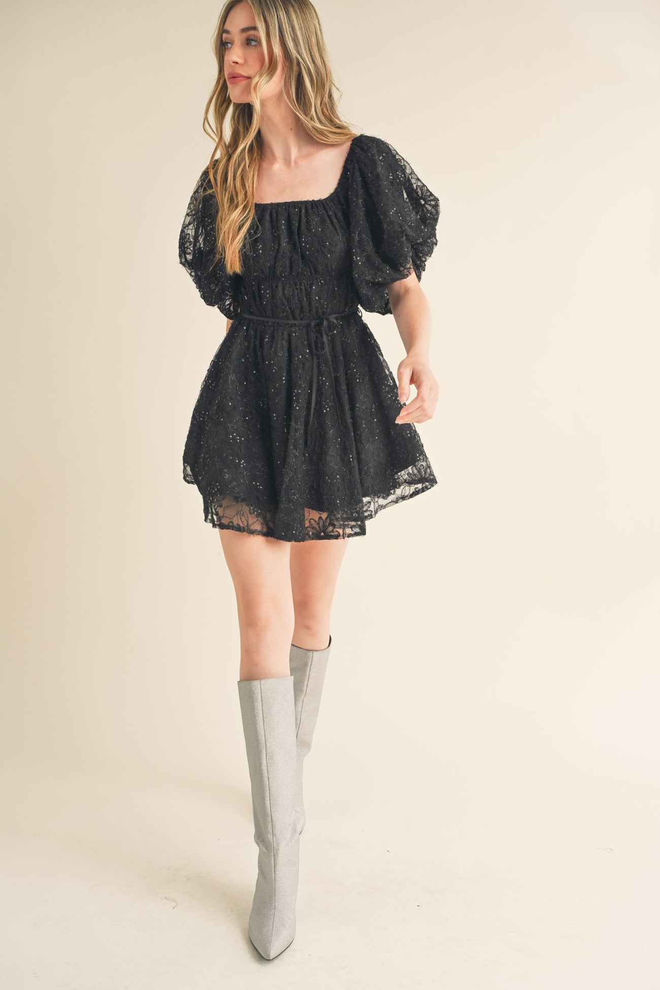 woman wearing a black lace puff sleeve mini dress with a cinched waist and silver knee-high boots