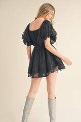 woman wearing a black short lace dress with puff sleeves and a fitted waist, showing a bare back and styled with silver knee-high boots