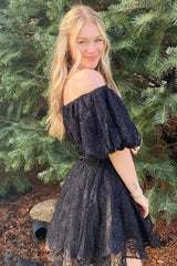 woman wearing a black off-the-shoulder dress with puff sleeves and a tiered skirt, standing outdoors among greenery