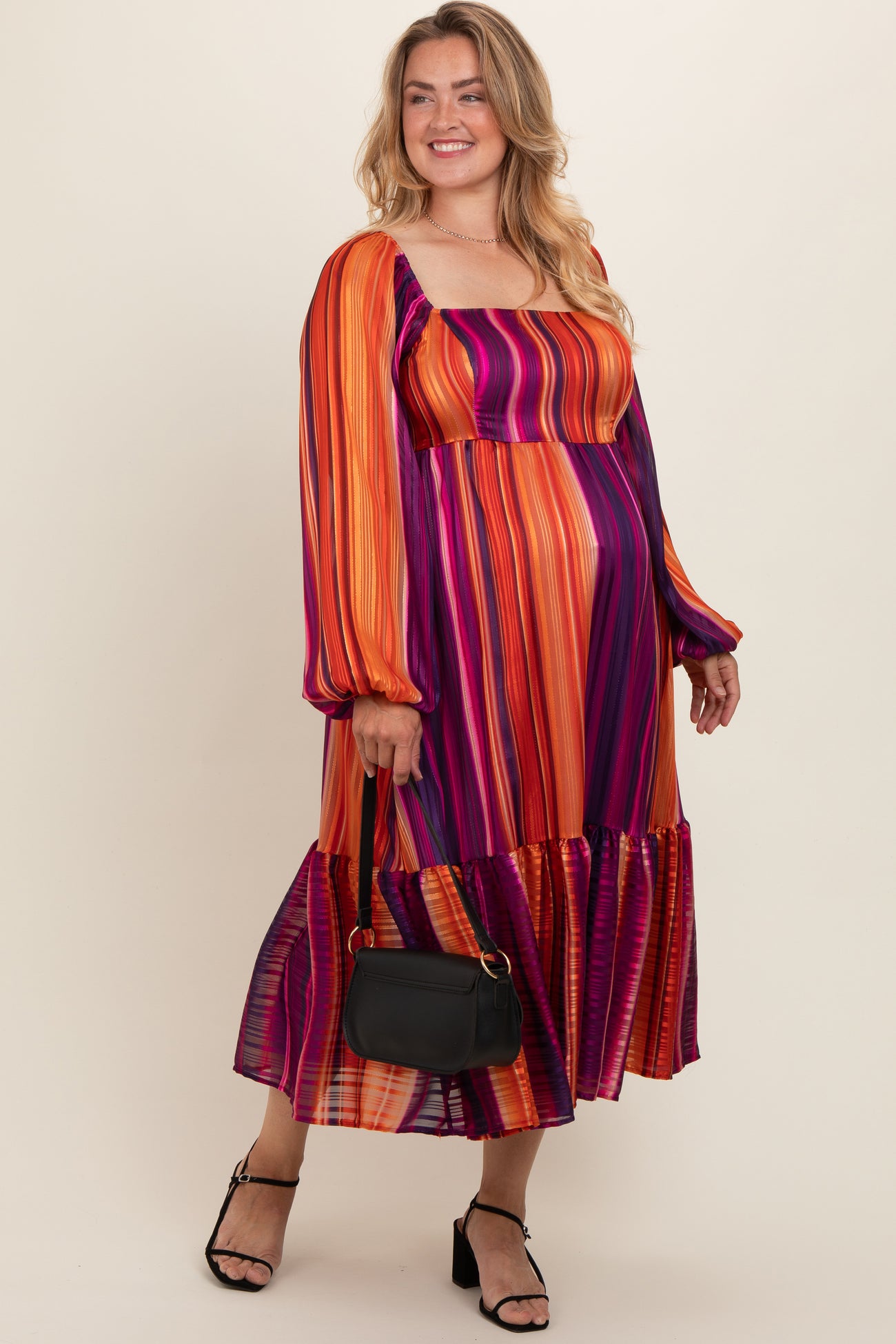 woman wearing a colorful striped maternity plus maxi dress with long sleeves and a flowing skirt, holding a black handbag
