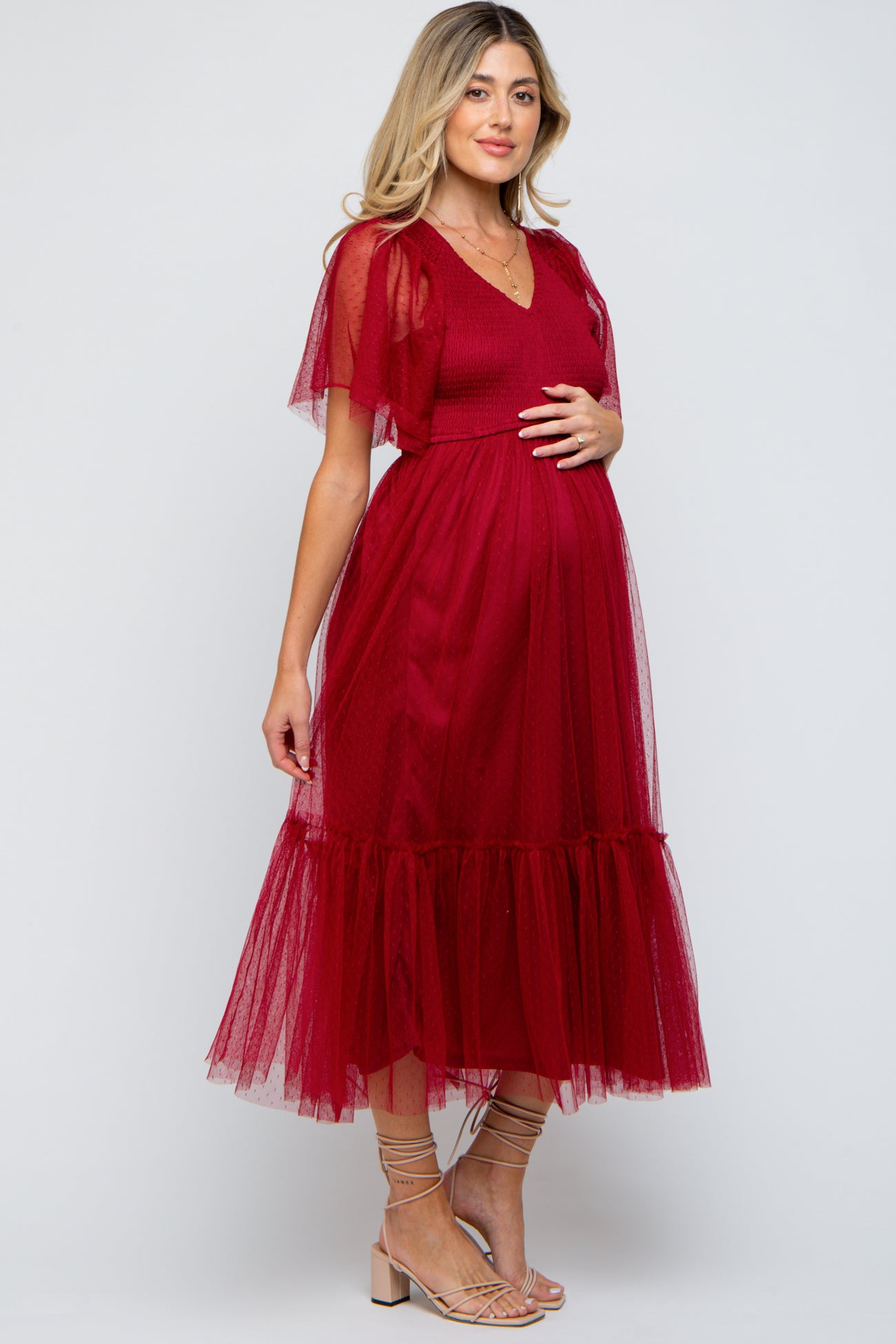 woman wearing a maroon maternity dress with sheer flutter sleeves and a tiered skirt, holding her baby bump