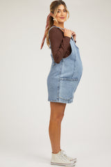 woman wearing a light blue denim maternity overall dress and a brown long sleeve top, showcasing her baby bump