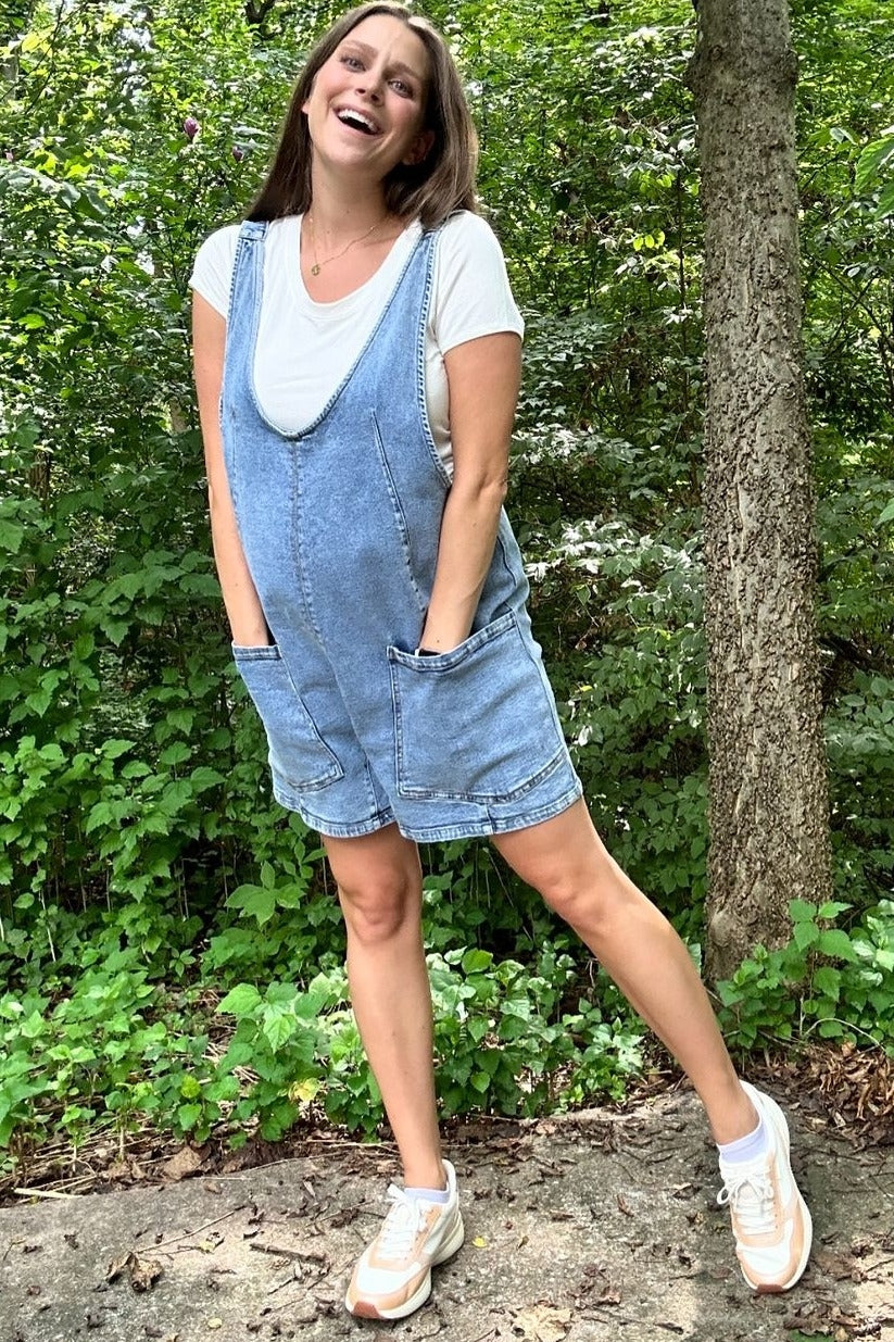 woman wearing a light blue denim maternity romper with pockets over a white t-shirt and styled with sneakers