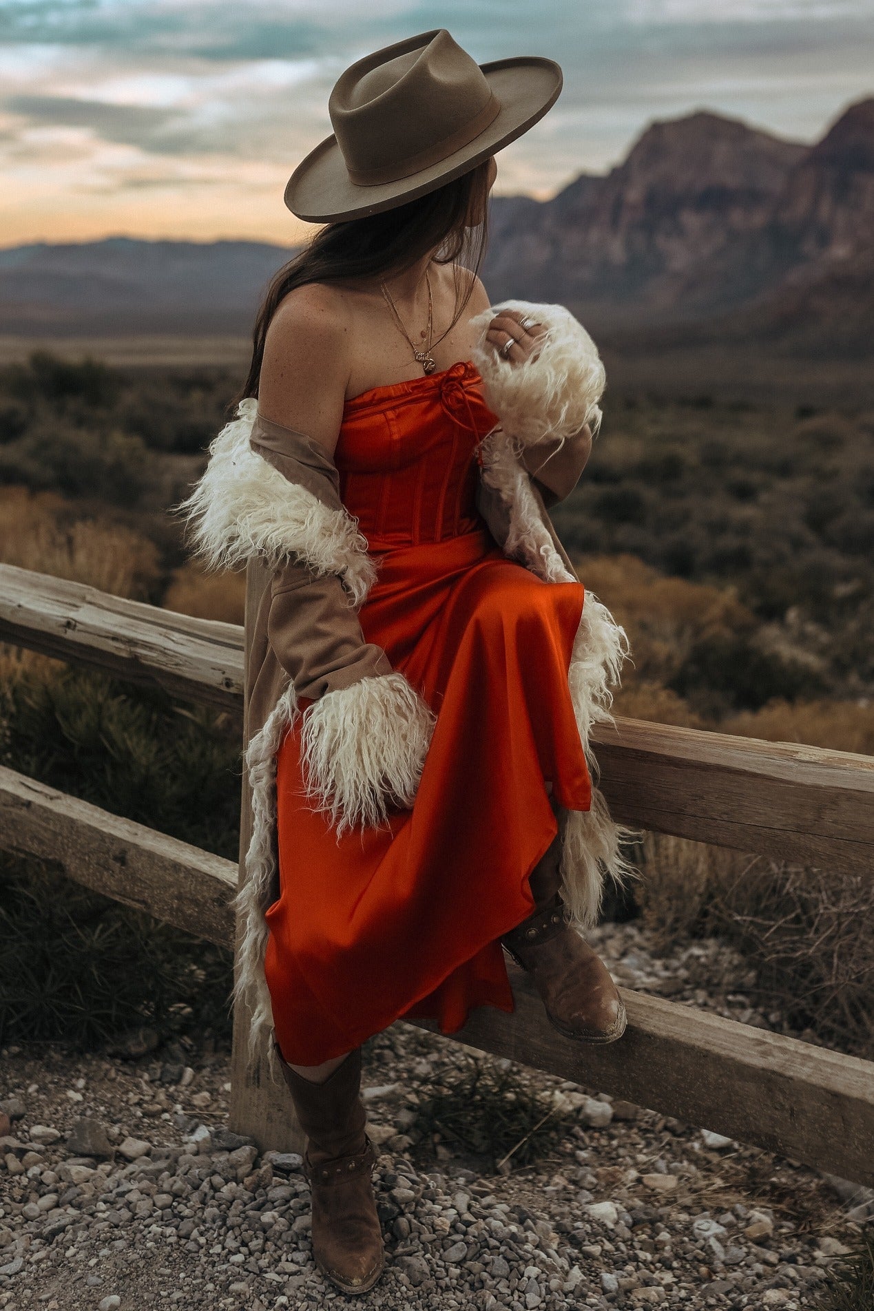 woman wearing a red strapless dress with a fur shawl and wide-brimmed hat, sitting on a wooden fence in a mountainous landscape