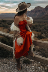 woman wearing a red strapless dress with a fur shawl and wide-brimmed hat, sitting on a wooden fence in a mountainous landscape