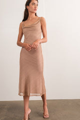 woman wearing a tan midi dress with a draped neckline and slim straps, standing in a relaxed pose