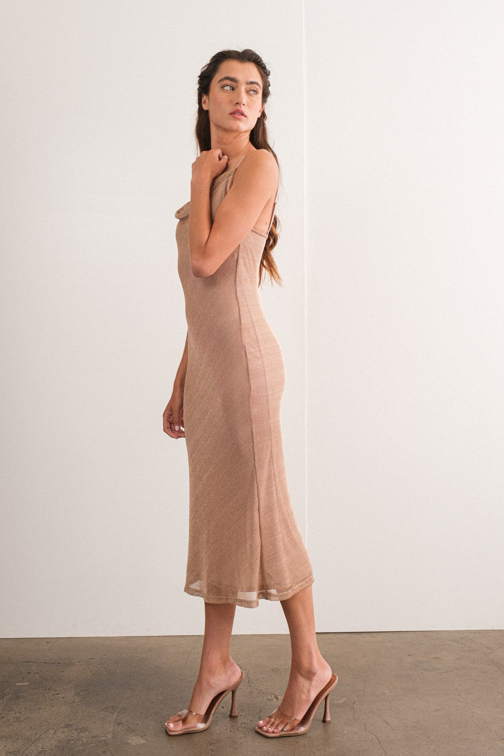 woman wearing a beige sleeveless midi dress and clear high-heeled sandals, posed sideways against a plain background