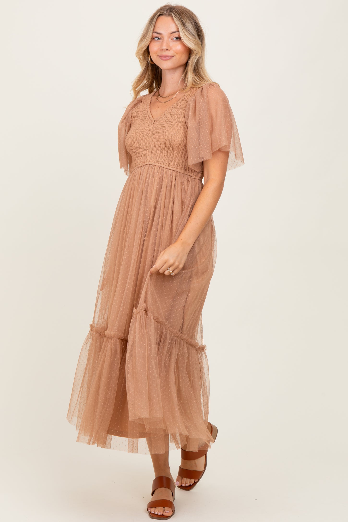 woman wearing a tan long sleeve tulle maxi dress with a smocked bodice and ruffled hem, standing on a light background