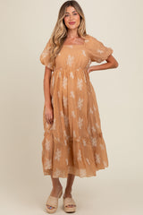 woman wearing a tan maternity dress with floral embroidery and short puff sleeves, standing and showing off her baby bump